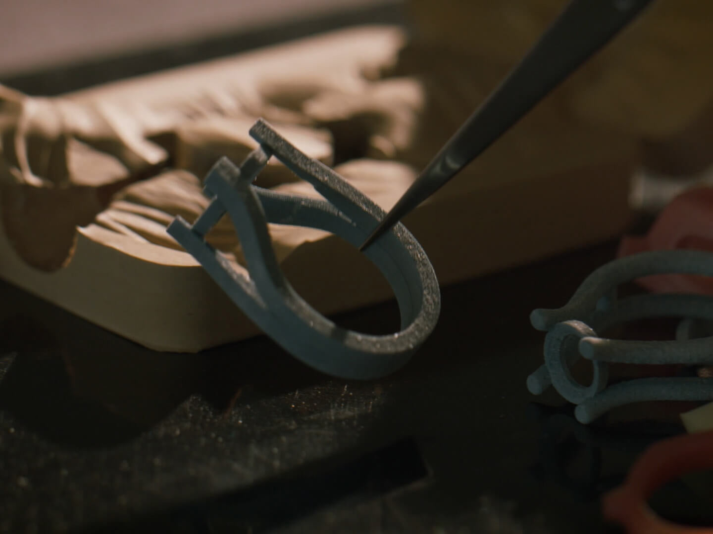 A close-up of a light blue 3D-printed ring-like component being held by tweezers over a dark work surface, with a blurred beige object in the background.