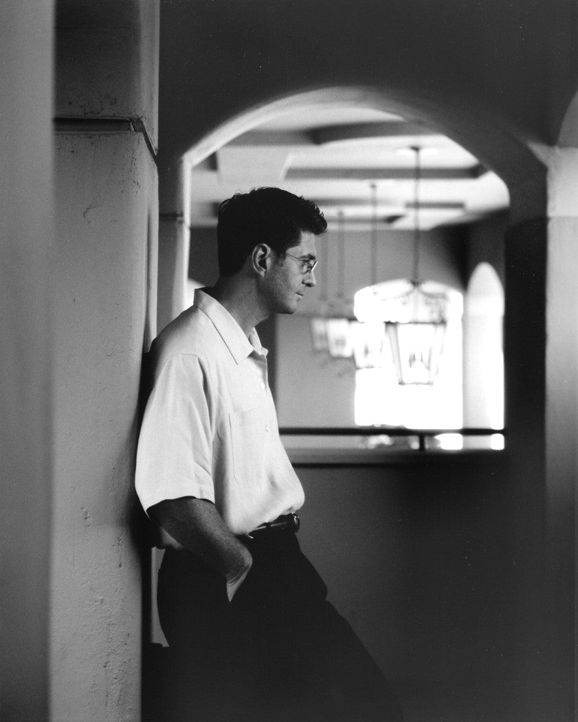 A portrait of Garo Demirjian leaning against a wall in a corridor.