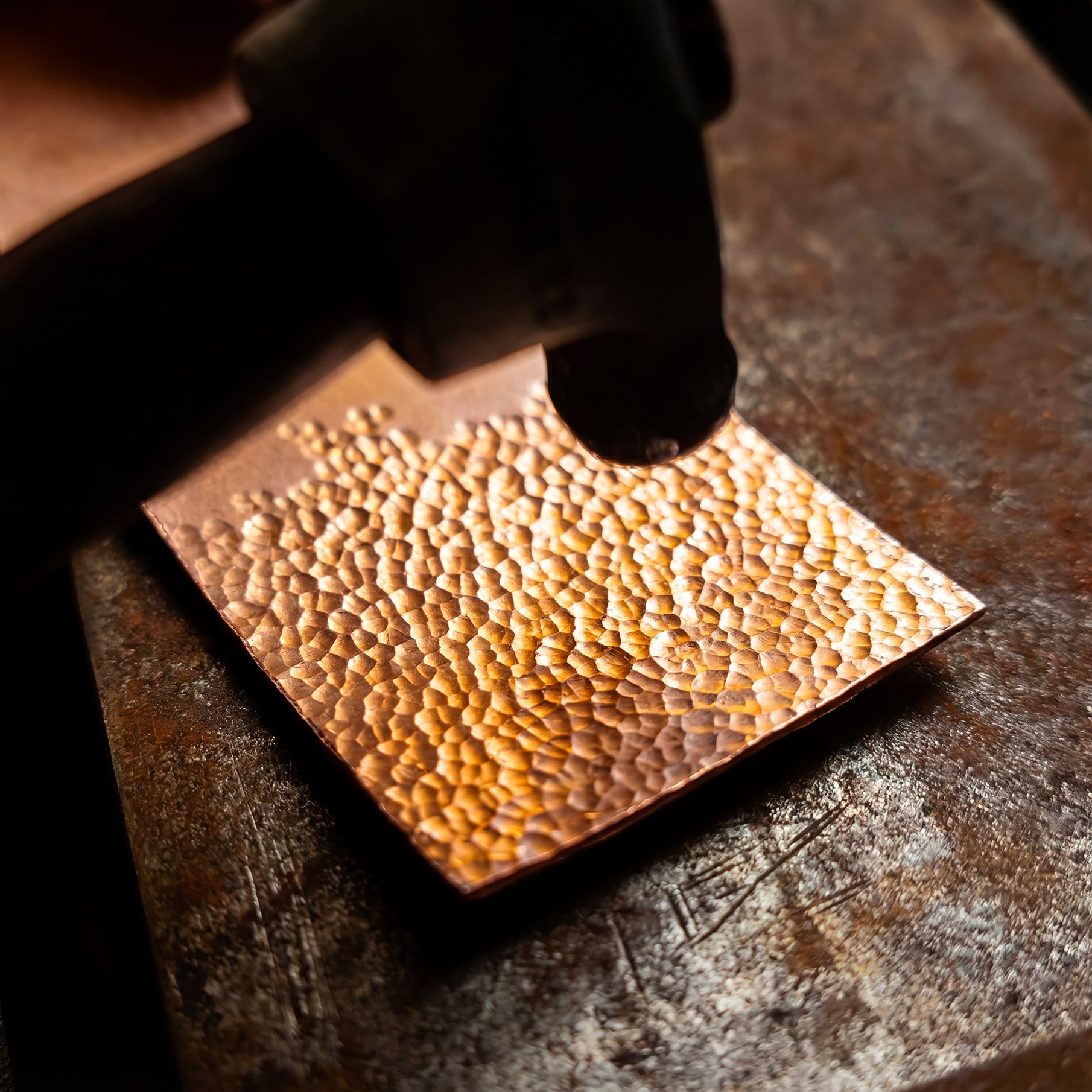An artisan metalworker striking a metal plate to create a decorative pattern, highlighting the craftsmanship of handmade jewelry.
