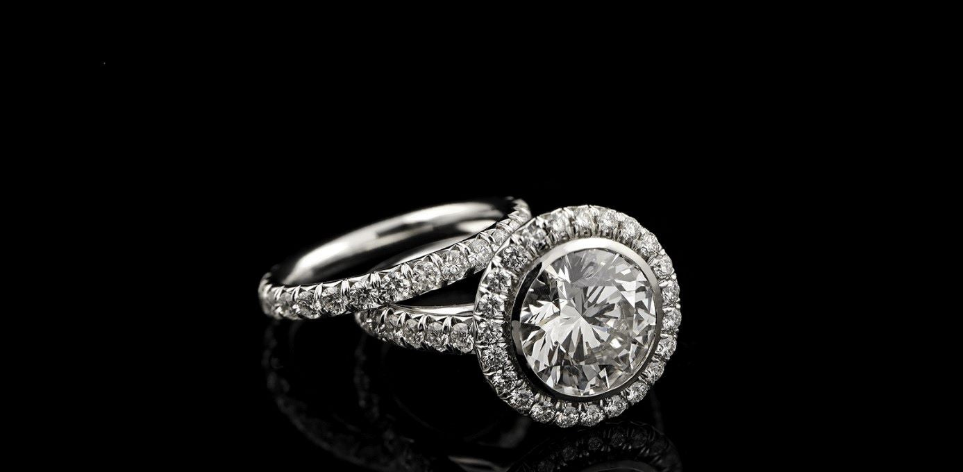 A close-up of a diamond ring on a black background. The ring is a custom-design by Garo Demirjian, diamond wedding ring with a solitaire diamond in the center. The diamond is surrounded by smaller diamonds on a pavé setting.