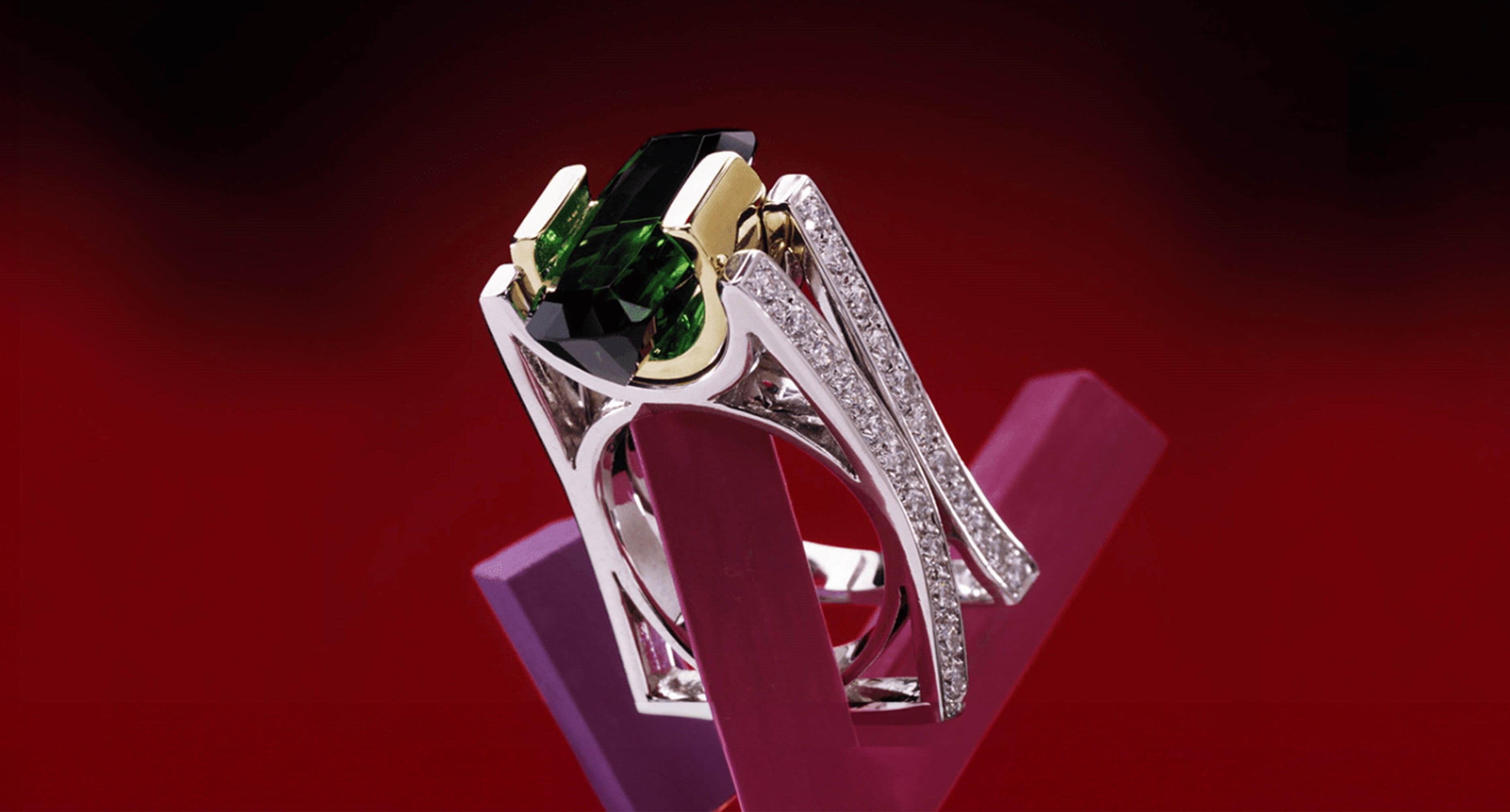 A custom-designed white gold ring with a prominent green emerald, showcased against a red background.
