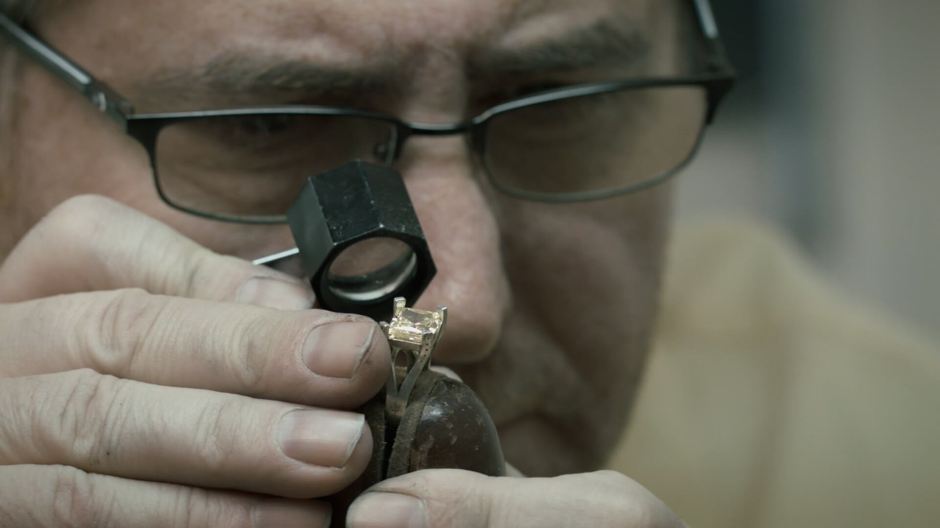 A close-up of a jeweler with glasses and a loupe inspecting a golden ring with a square-cut gemstone, holding it securely with a metal tool.