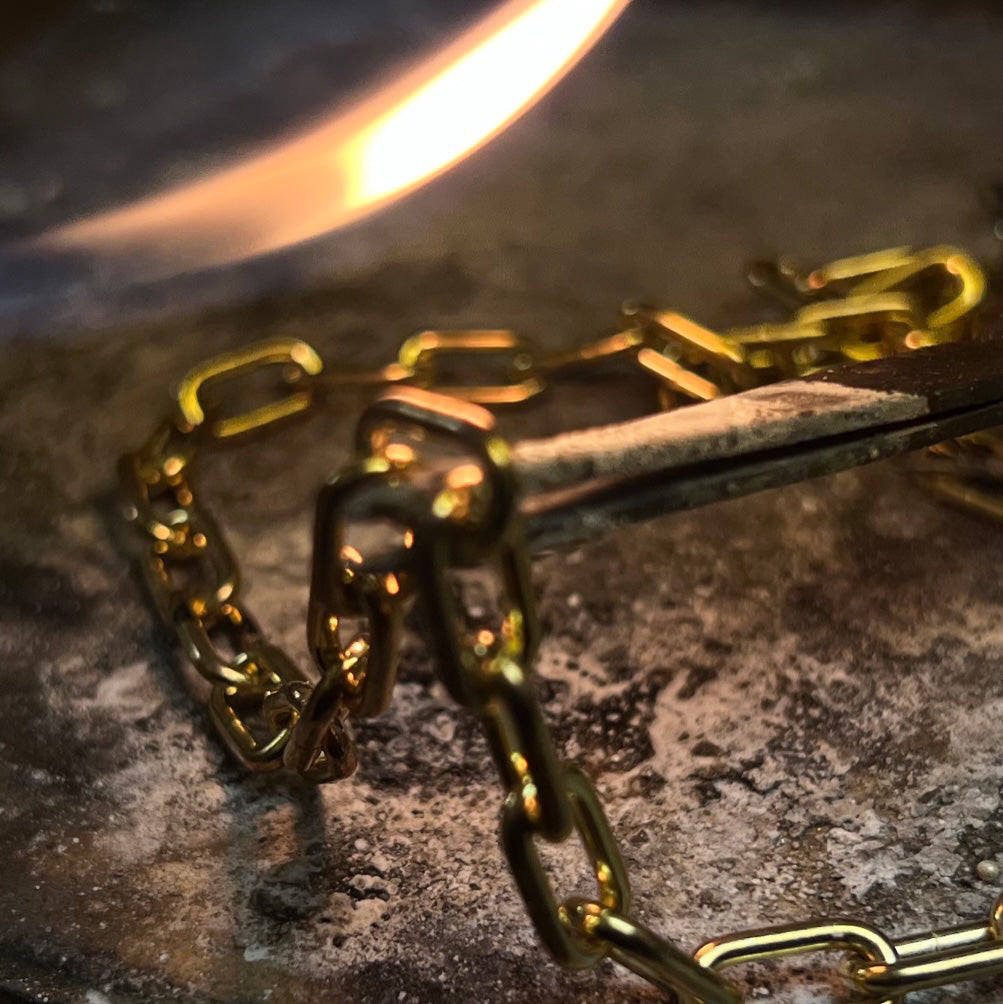A close-up of a gold chain being soldered with a torch on a table. The chain is being repaired by a jeweler.