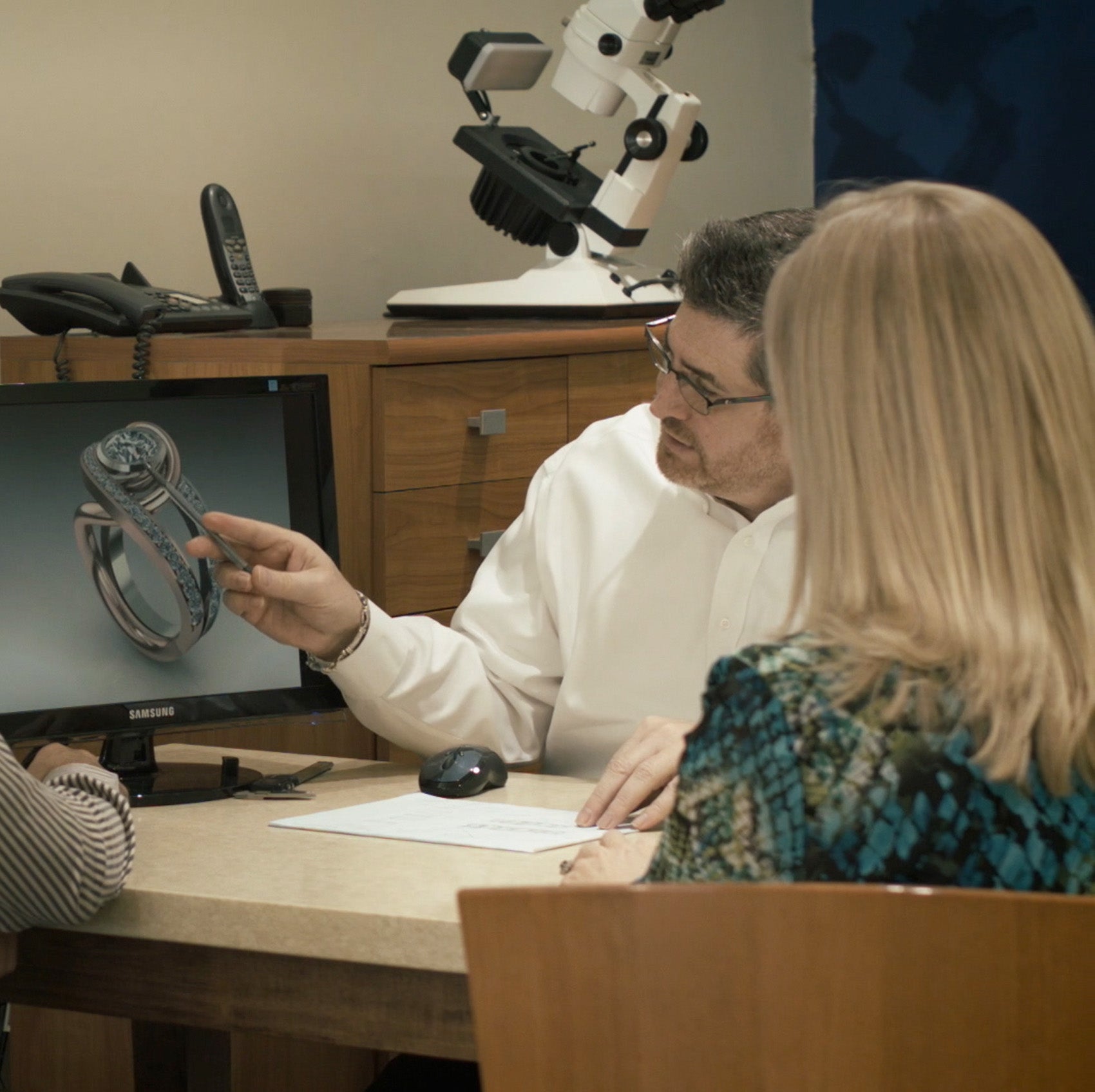 A jeweler shows a 3D computer rendering of a custom diamond ring design to a couple during a consultation appointment.