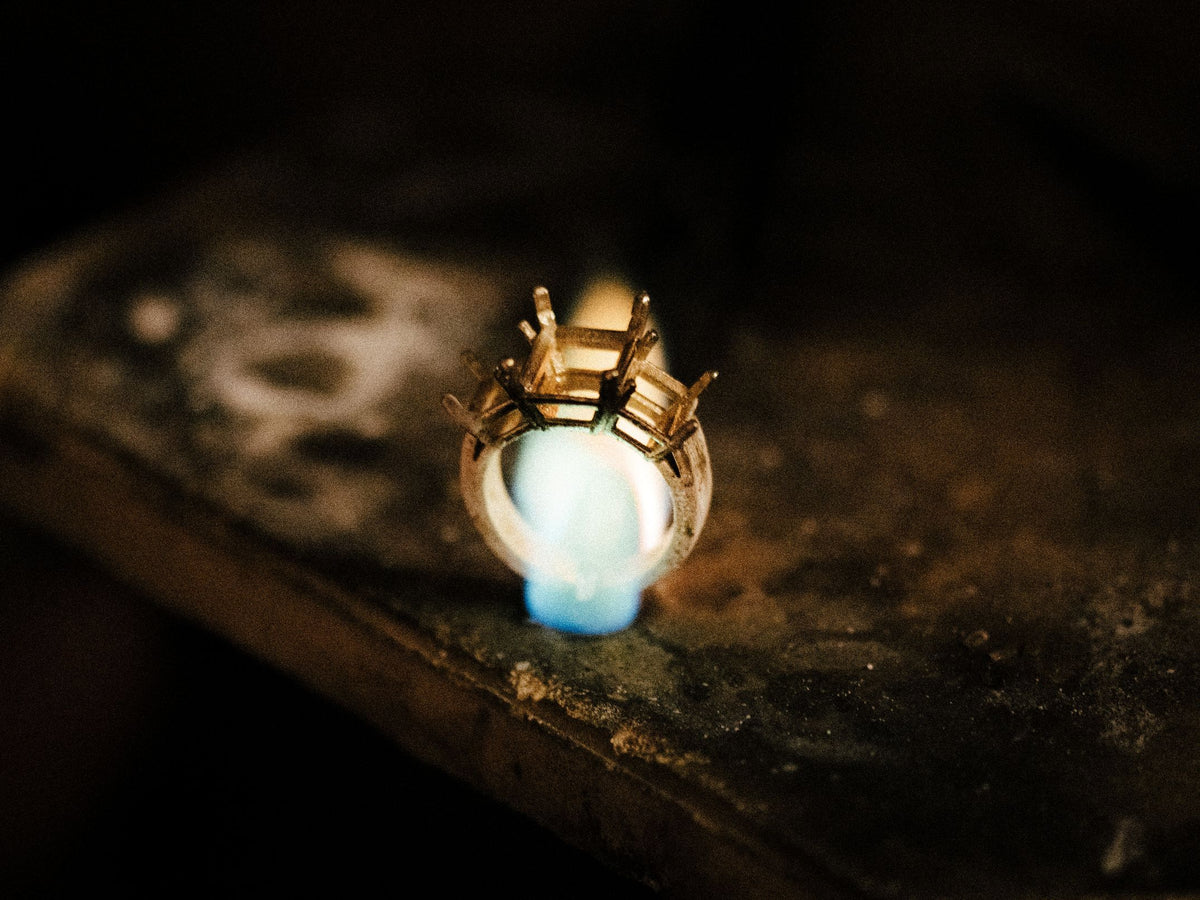 Close-up of a gold ring setting being heated by a blue torch flame on a jeweler's workbench during the manufacturing process.