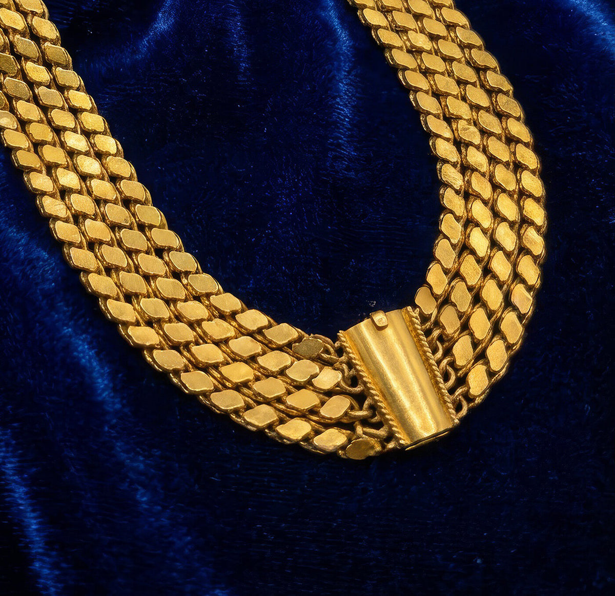 Close up of a vintage 22K gold jewelry: a heavy yellow gold triple-strand necklace with a wide woven design and barrel clasp resting on a dark blue fabric surface.