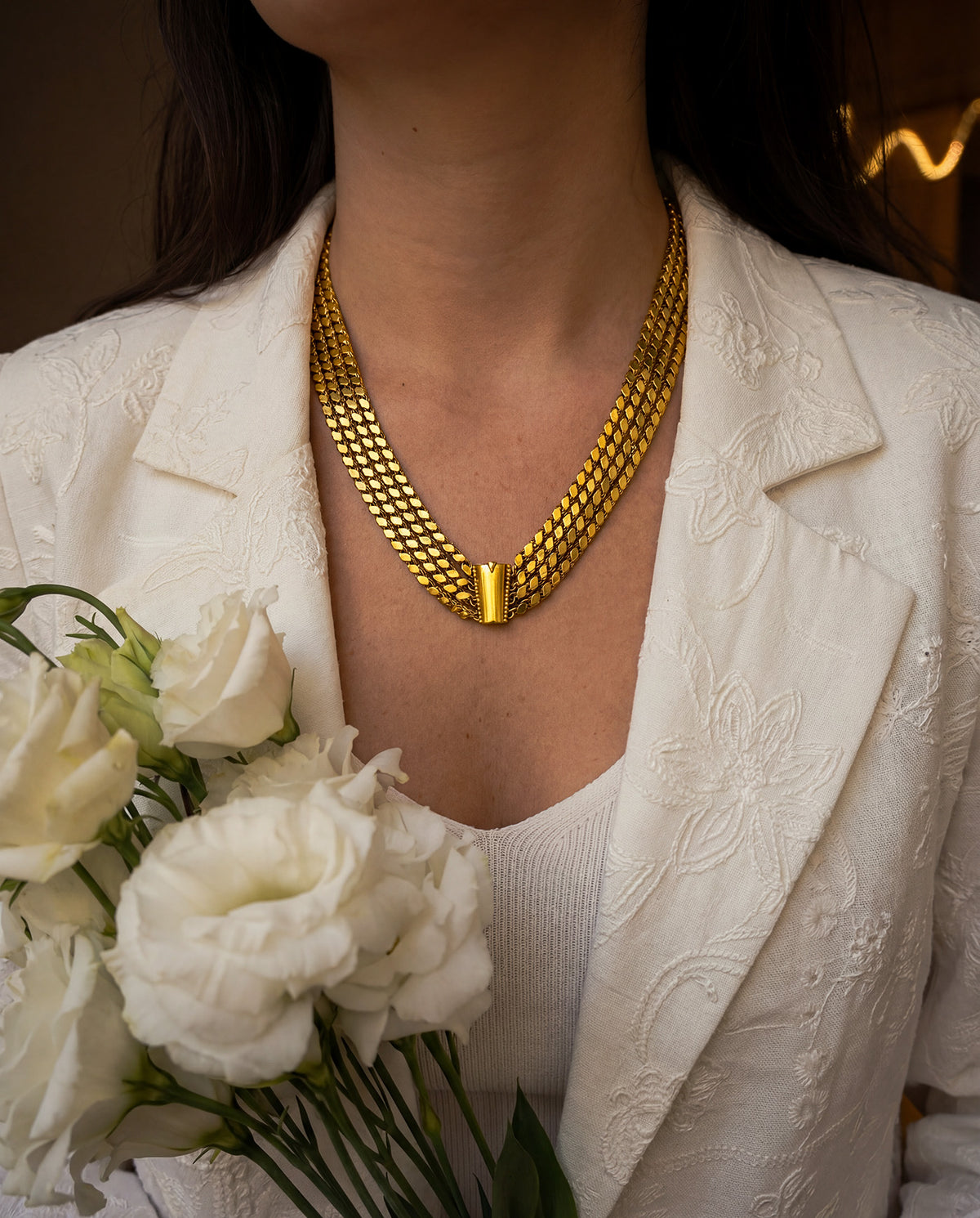 A woman wearing a vintage 22K yellow gold wide triple strand necklace with a white blazer, holding white flowers.